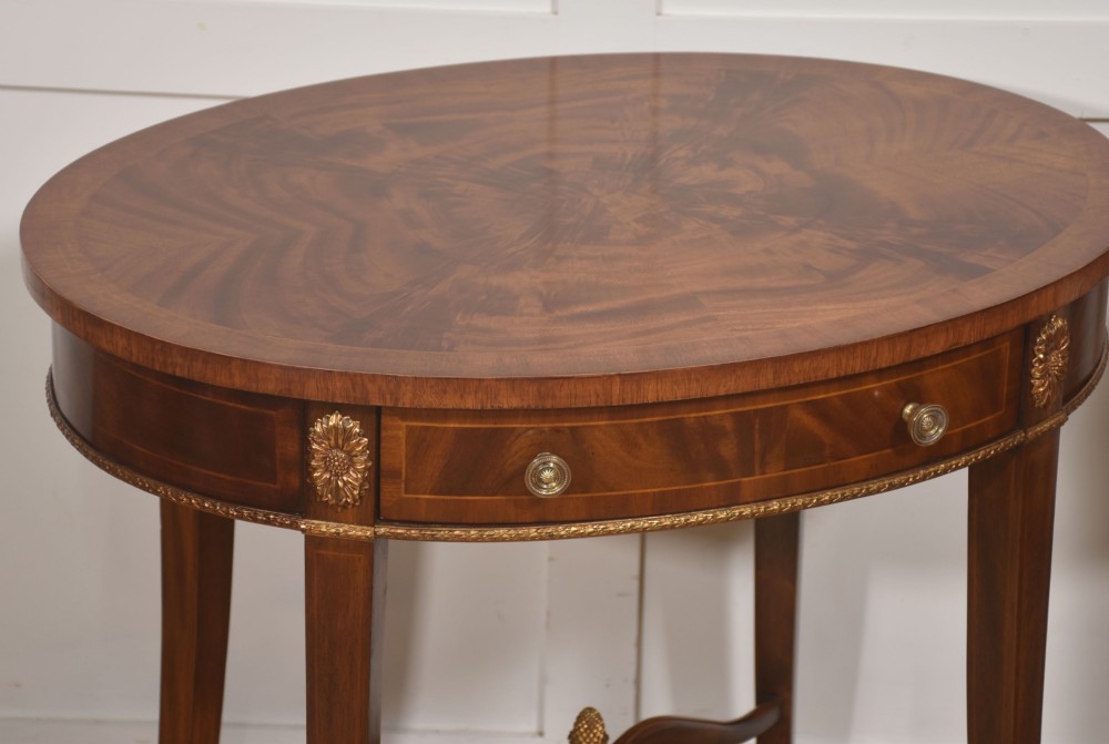 A pair of oval flame mahogany side tables featuring brass rosette mounts, single drawers, curved legs, and an X-shaped stretcher with a brass finial. View VI