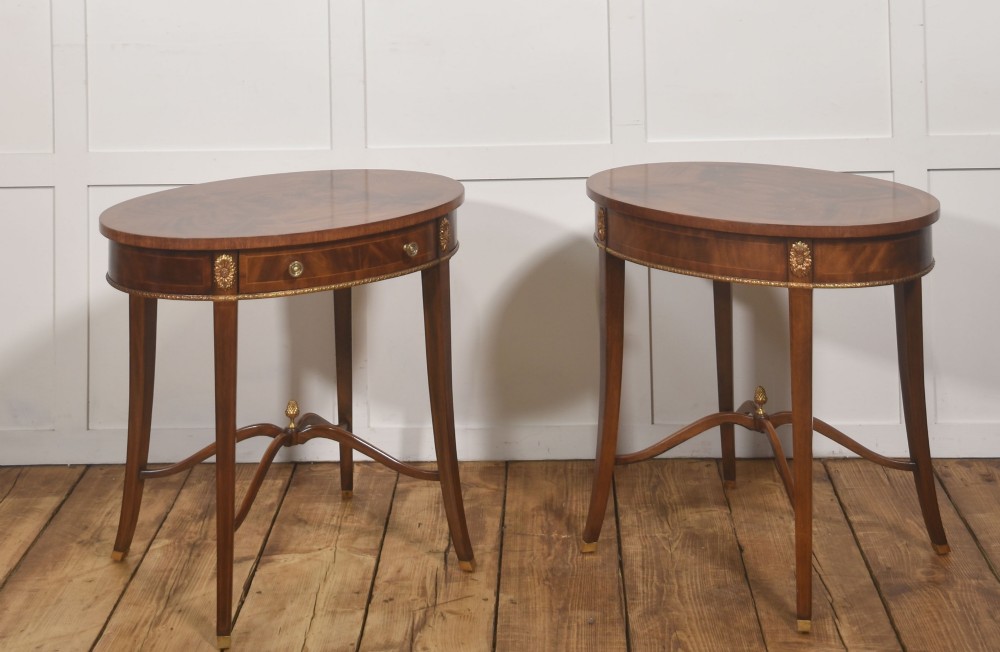 A pair of oval flame mahogany side tables featuring brass rosette mounts, single drawers, curved legs, and an X-shaped stretcher with a brass finial. View IX