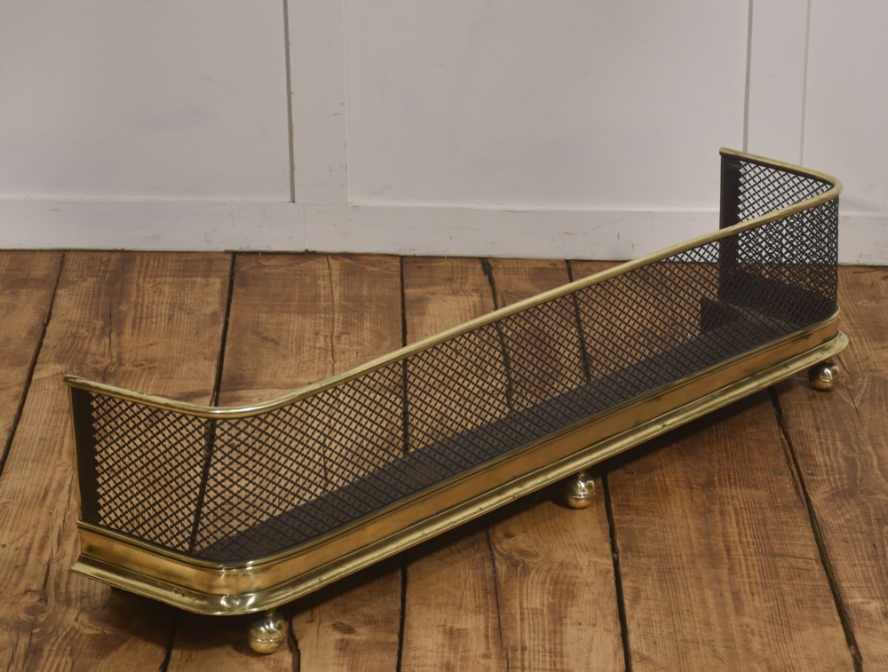 A low rectangular fireplace fender featuring a polished brass top rim and base moulding with black steel diamond mesh sides on small brass ball feet.