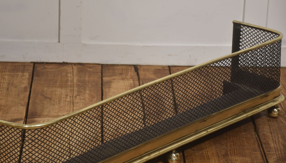A low rectangular fireplace fender featuring a polished brass top rim and base moulding with black steel diamond mesh sides on small brass ball feet. View III