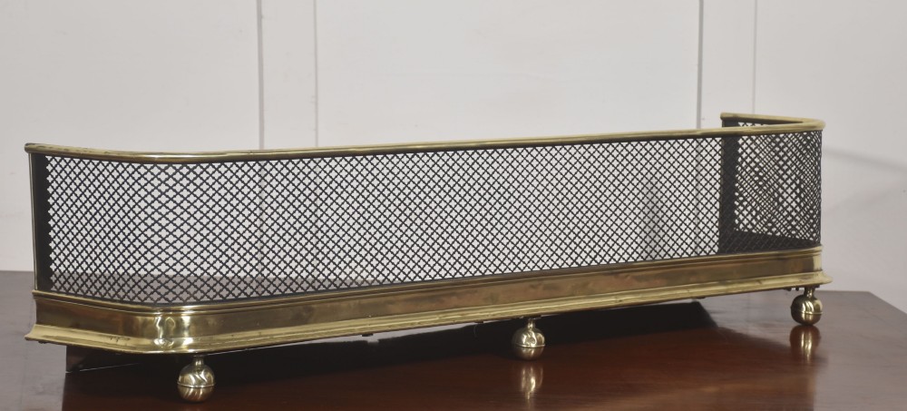 A low rectangular fireplace fender featuring a polished brass top rim and base moulding with black steel diamond mesh sides on small brass ball feet. View IV