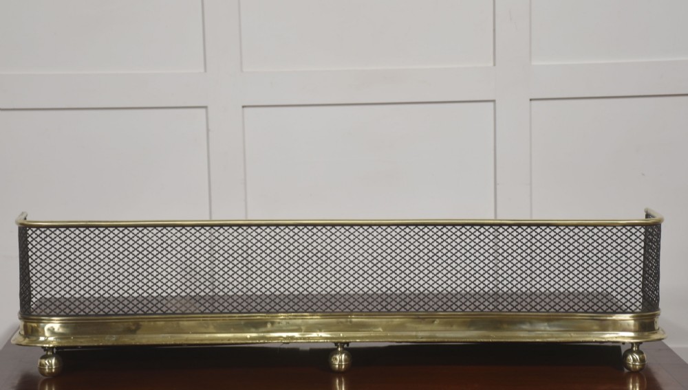 A low rectangular fireplace fender featuring a polished brass top rim and base moulding with black steel diamond mesh sides on small brass ball feet. View V