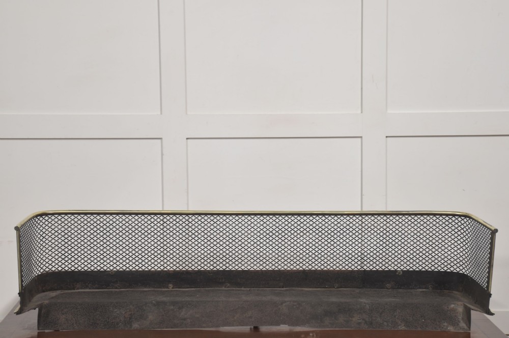 A low rectangular fireplace fender featuring a polished brass top rim and base moulding with black steel diamond mesh sides on small brass ball feet. View VII