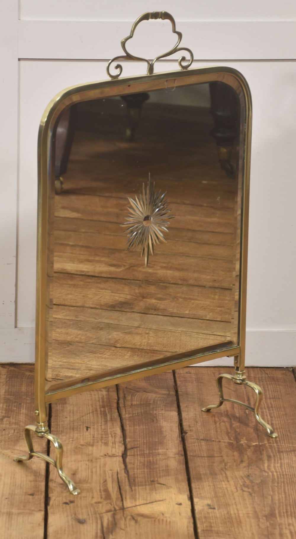 An arched brass-framed fire screen featuring a central cut-glass starburst pattern on a bevelled mirrored panel with a decorative top handle and splayed feet.