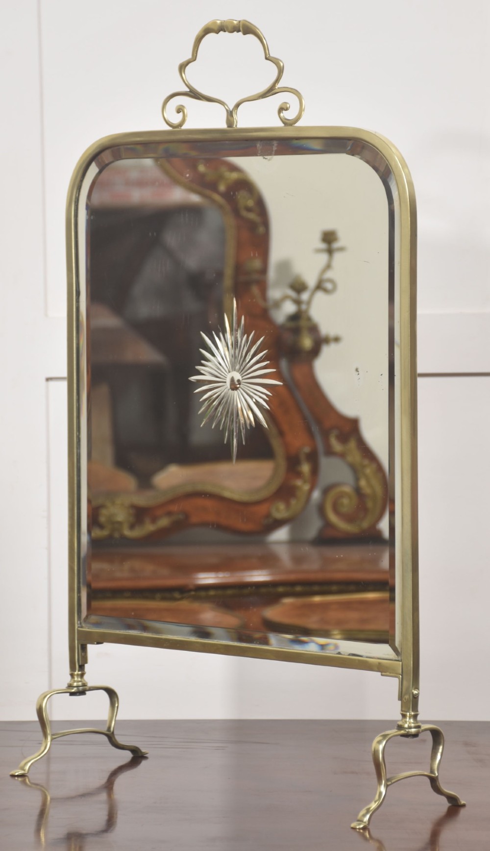 An arched brass-framed fire screen featuring a central cut-glass starburst pattern on a bevelled mirrored panel with a decorative top handle and splayed feet. View V
