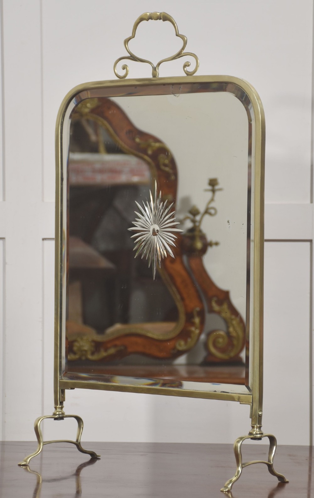 An arched brass-framed fire screen featuring a central cut-glass starburst pattern on a bevelled mirrored panel with a decorative top handle and splayed feet. View VIII