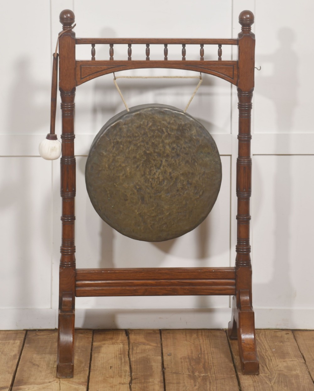 A standing dark oak frame featuring turned upright pillars, top spindles, and a central suspended hammered brass dinner gong dish with a hanging mallet.