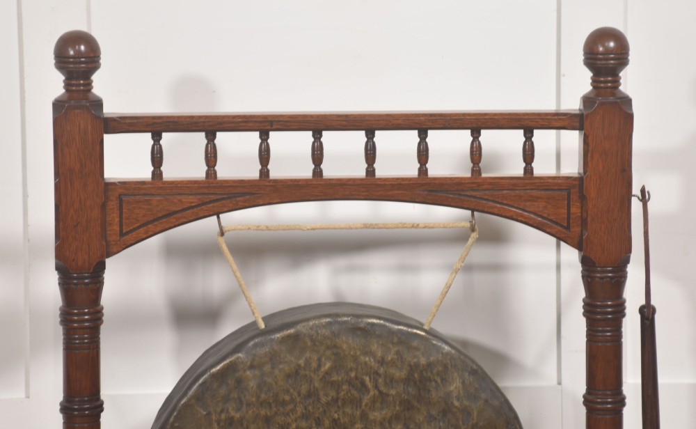 A standing dark oak frame featuring turned upright pillars, top spindles, and a central suspended hammered brass dinner gong dish with a hanging mallet. View III
