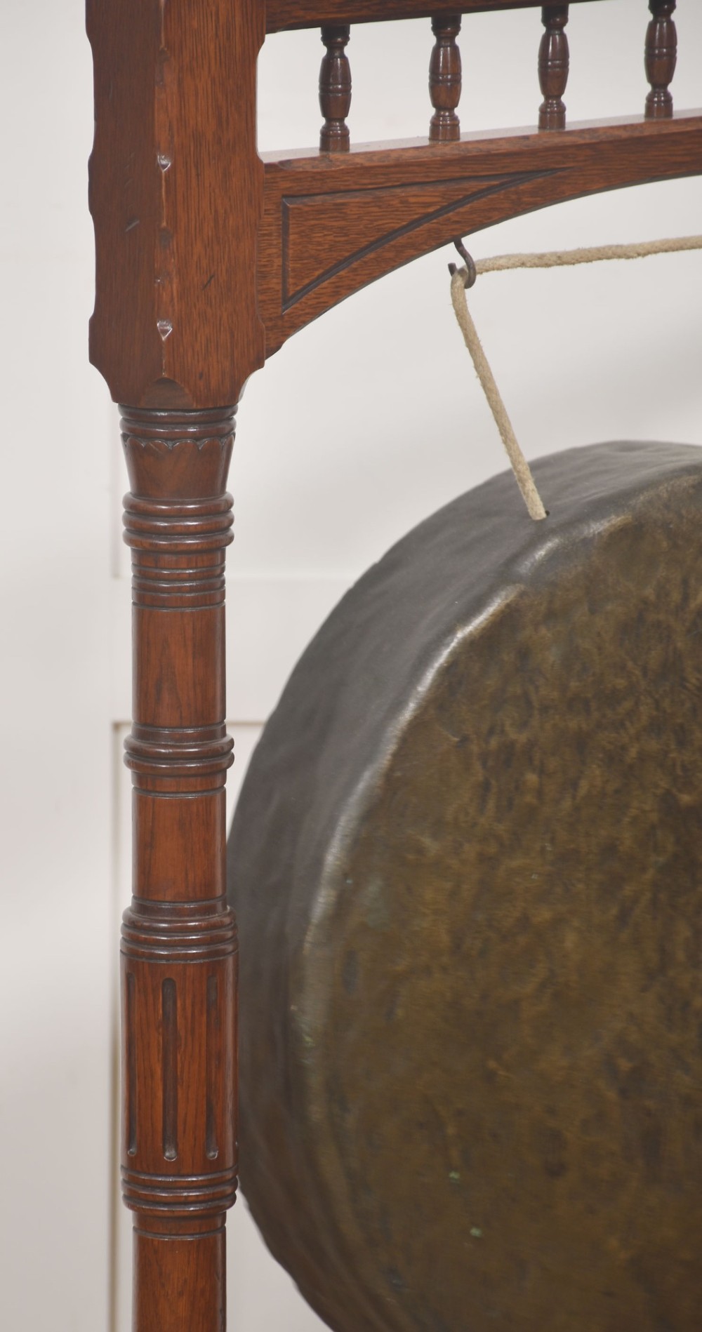 A standing dark oak frame featuring turned upright pillars, top spindles, and a central suspended hammered brass dinner gong dish with a hanging mallet. View V