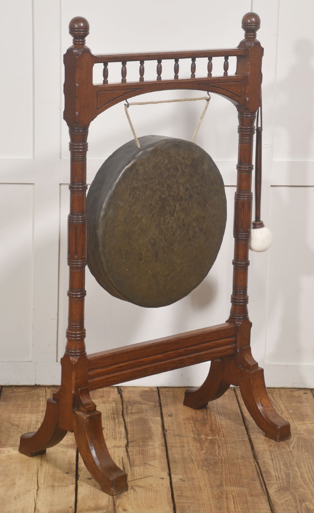 A standing dark oak frame featuring turned upright pillars, top spindles, and a central suspended hammered brass dinner gong dish with a hanging mallet. View VI