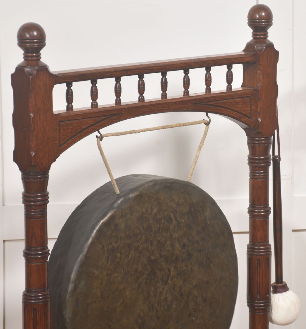 A standing dark oak frame featuring turned upright pillars, top spindles, and a central suspended hammered brass dinner gong dish with a hanging mallet. View VII