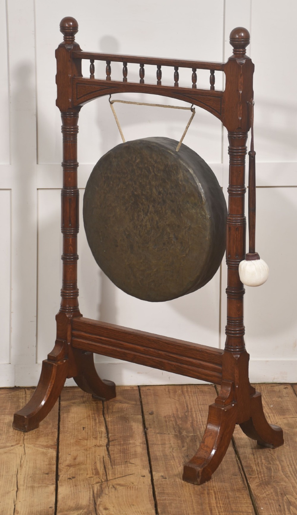 A standing dark oak frame featuring turned upright pillars, top spindles, and a central suspended hammered brass dinner gong dish with a hanging mallet. View IX