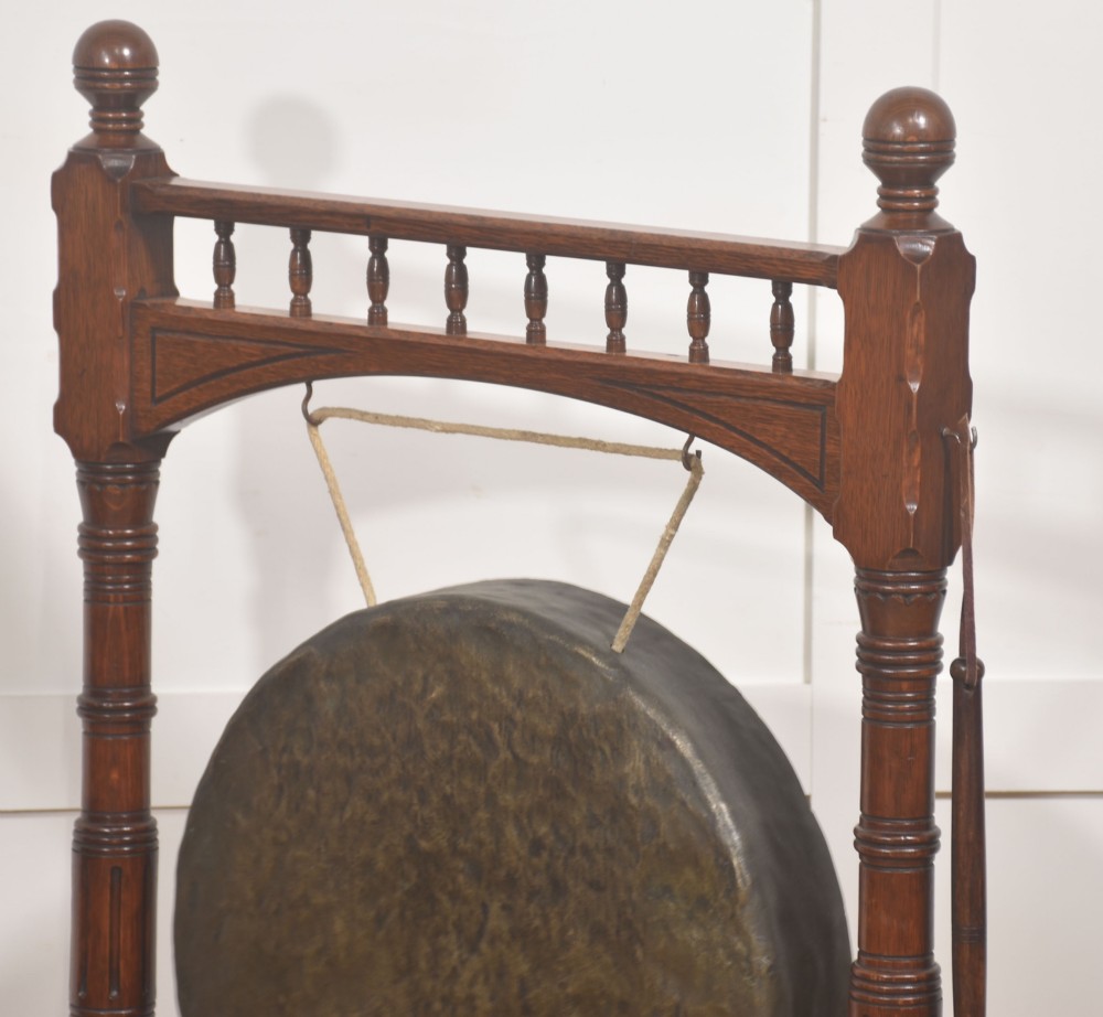 A standing dark oak frame featuring turned upright pillars, top spindles, and a central suspended hammered brass dinner gong dish with a hanging mallet. View X