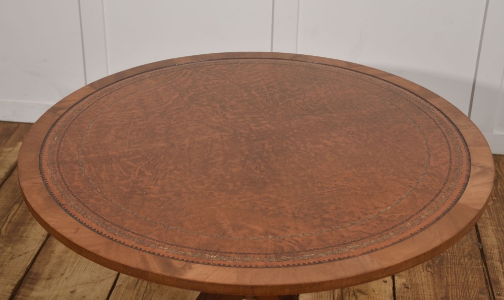A circular Regency style mahogany coffee table featuring an inset brown leather top with gold leaf tooling, supported by a central pedestal and four splayed legs fitted with brass castors. View V