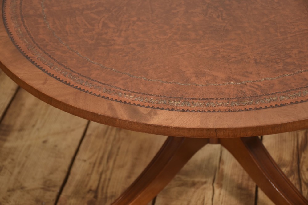 A circular Regency style mahogany coffee table featuring an inset brown leather top with gold leaf tooling, supported by a central pedestal and four splayed legs fitted with brass castors. View VI