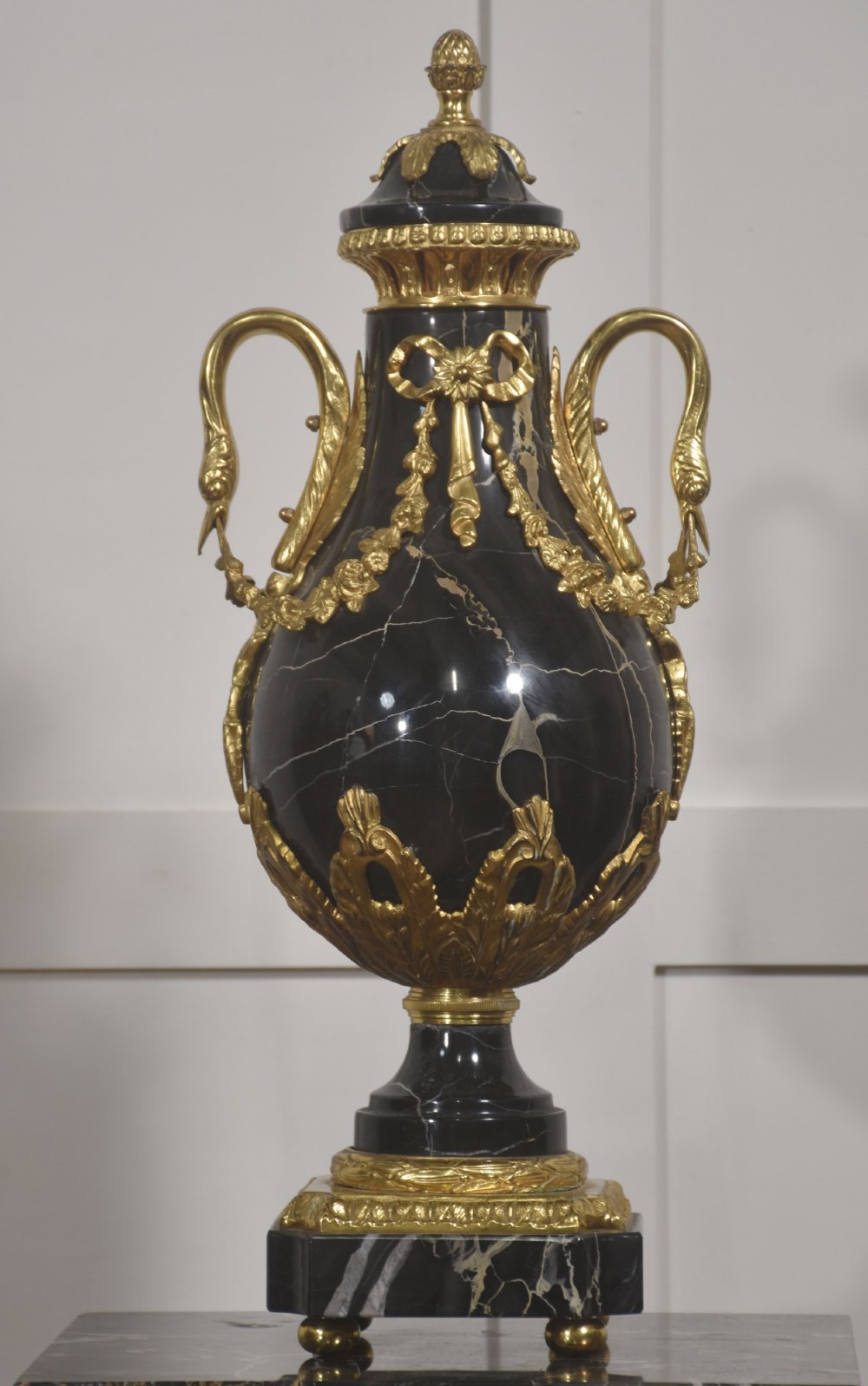 A black veined-marble urn with two handles and gilt-metal mounts resting on a matching cylindrical black marble pedestal with a Corinthian capital. View II