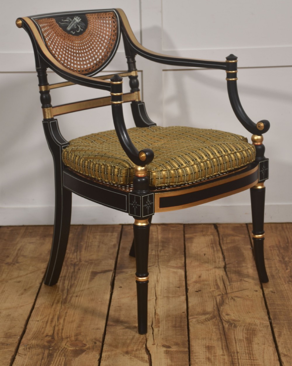 A Regency style ebonised and parcel-gilt open armchair featuring a curved cane backrest with painted medallion detail, sweeping arms, and a patterned gold upholstered cushion.