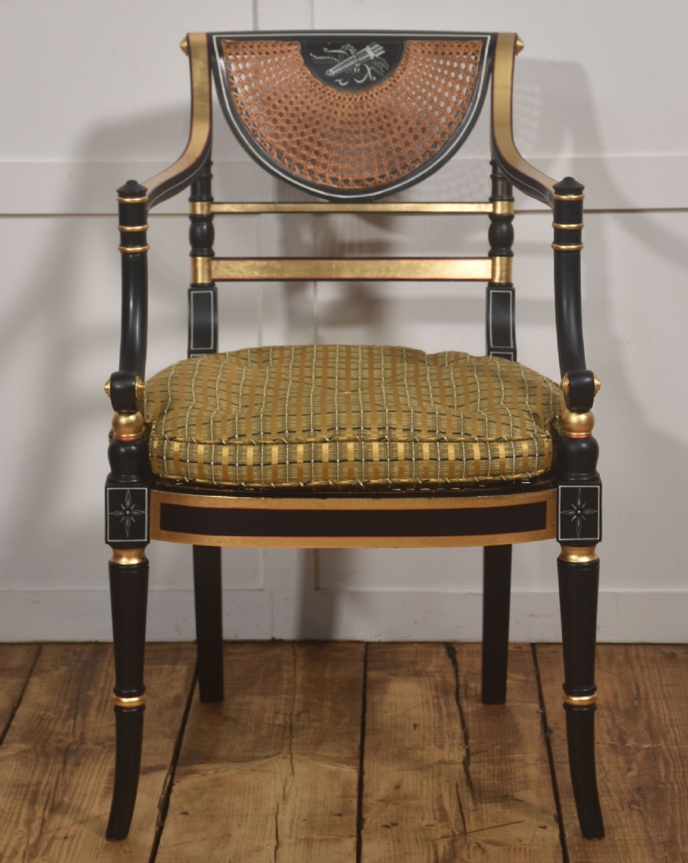 A Regency style ebonised and parcel-gilt open armchair featuring a curved cane backrest with painted medallion detail, sweeping arms, and a patterned gold upholstered cushion. View V