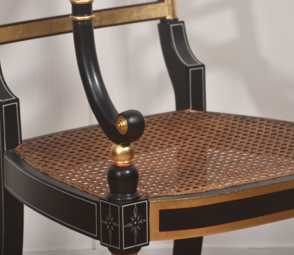 A Regency style ebonised and parcel-gilt open armchair featuring a curved cane backrest with painted medallion detail, sweeping arms, and a patterned gold upholstered cushion. View VI