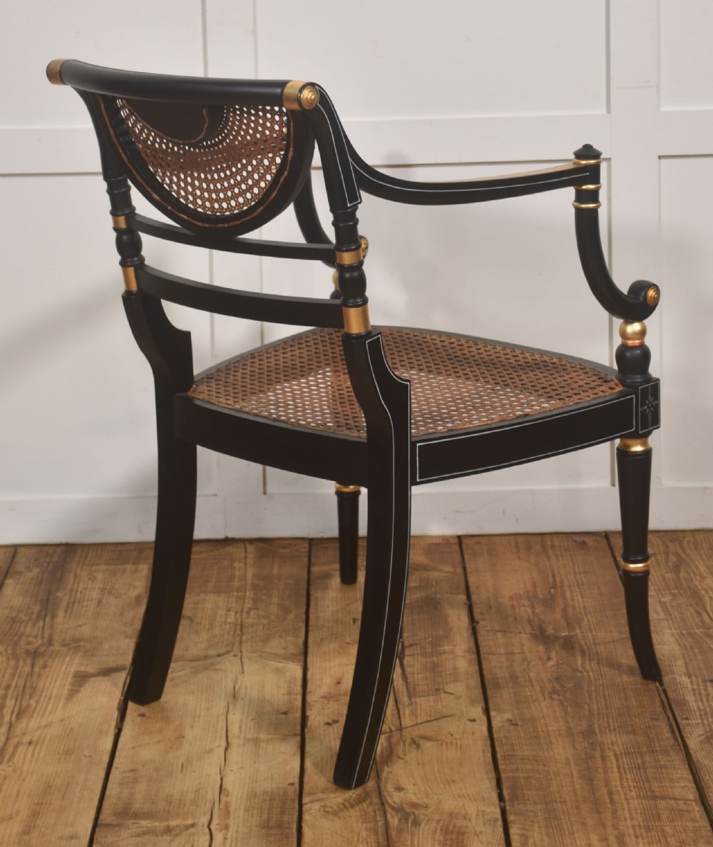 A Regency style ebonised and parcel-gilt open armchair featuring a curved cane backrest with painted medallion detail, sweeping arms, and a patterned gold upholstered cushion. View IX