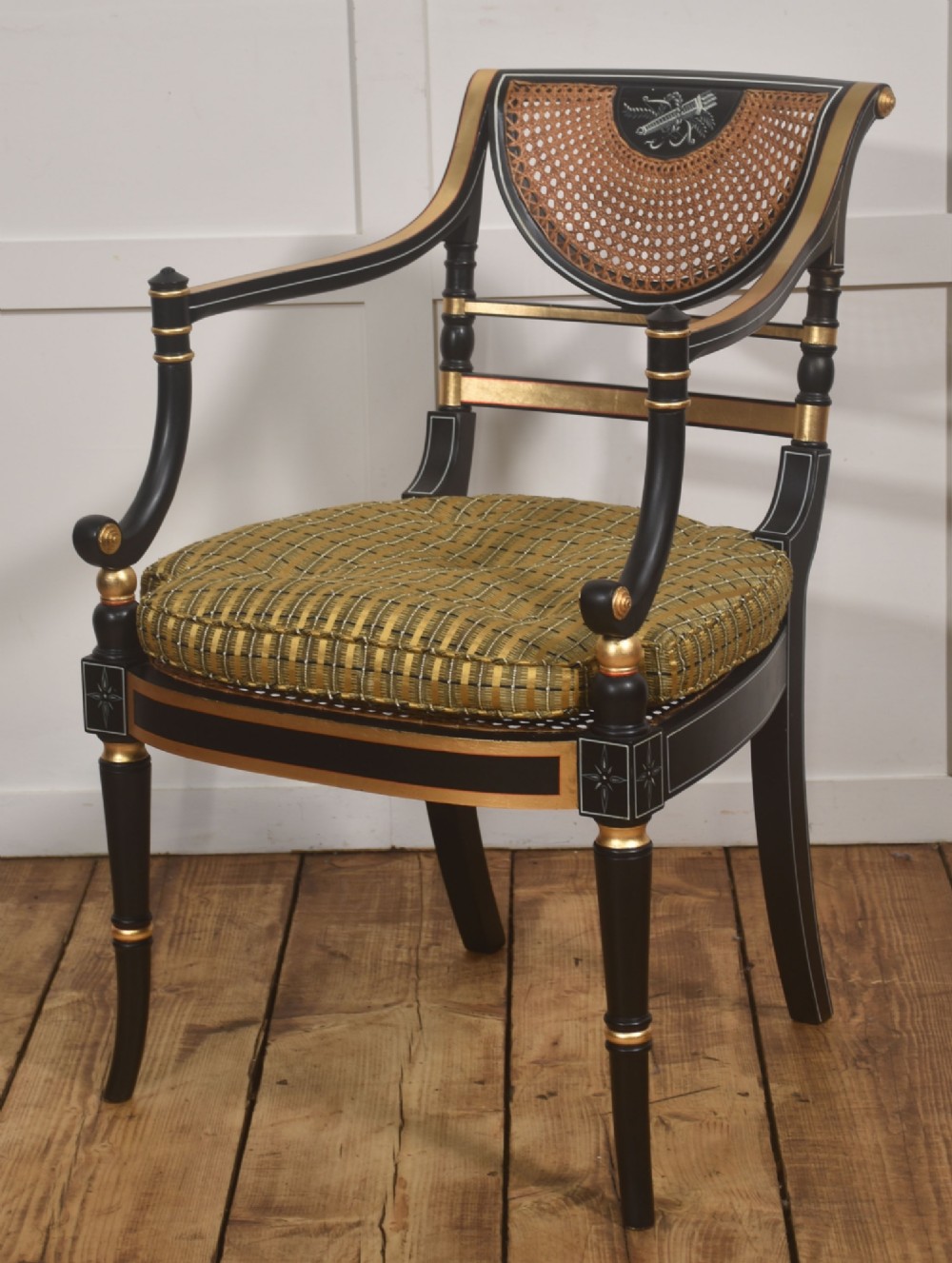 A Regency style ebonised and parcel-gilt open armchair featuring a curved cane backrest with painted medallion detail, sweeping arms, and a patterned gold upholstered cushion. View X