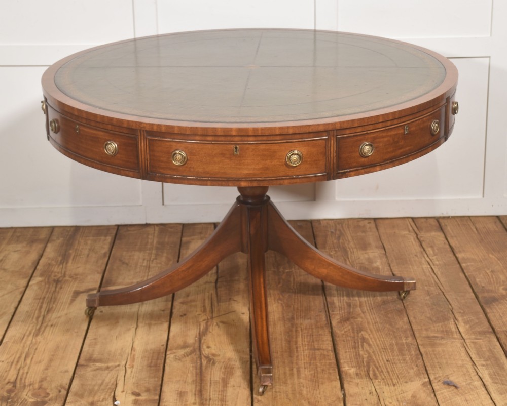 A circular mahogany drum table featuring a green leather top with gold tooling, multiple drawers with brass ring handles, and a four-legged pedestal base on castors.