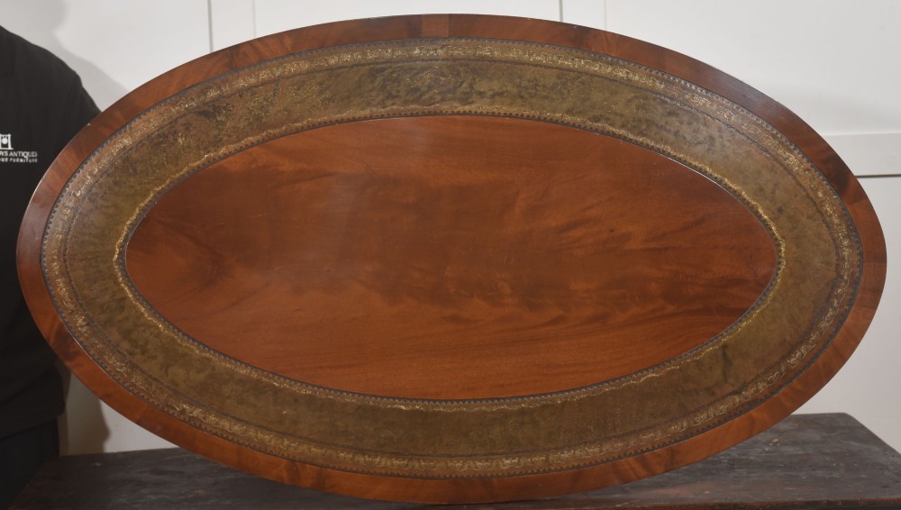 An oval mahogany coffee table featuring an inset green tooled leather border, a central turned pedestal stem, four swept legs, and brass castors. View II