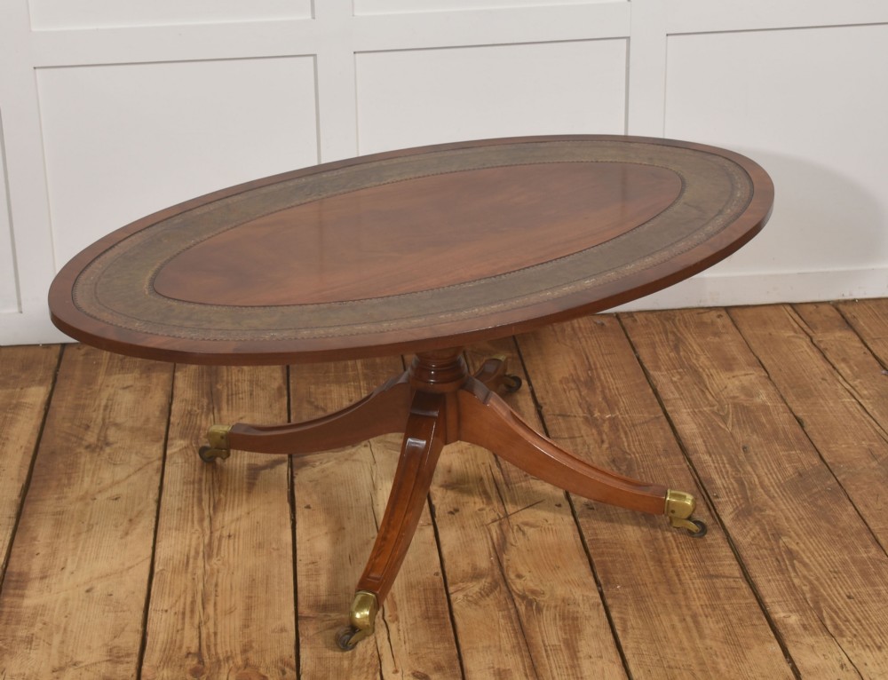 An oval mahogany coffee table featuring an inset green tooled leather border, a central turned pedestal stem, four swept legs, and brass castors.