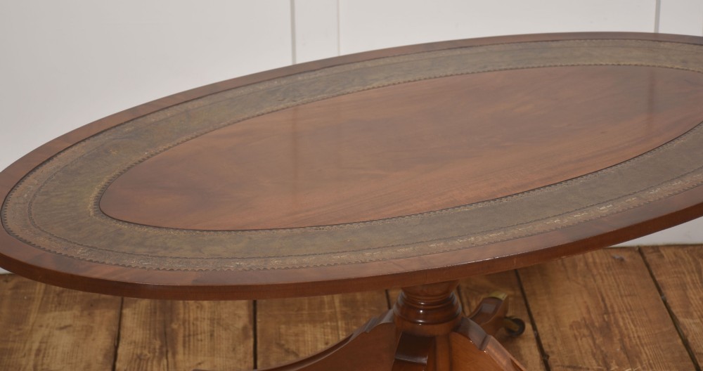 An oval mahogany coffee table featuring an inset green tooled leather border, a central turned pedestal stem, four swept legs, and brass castors. View IV