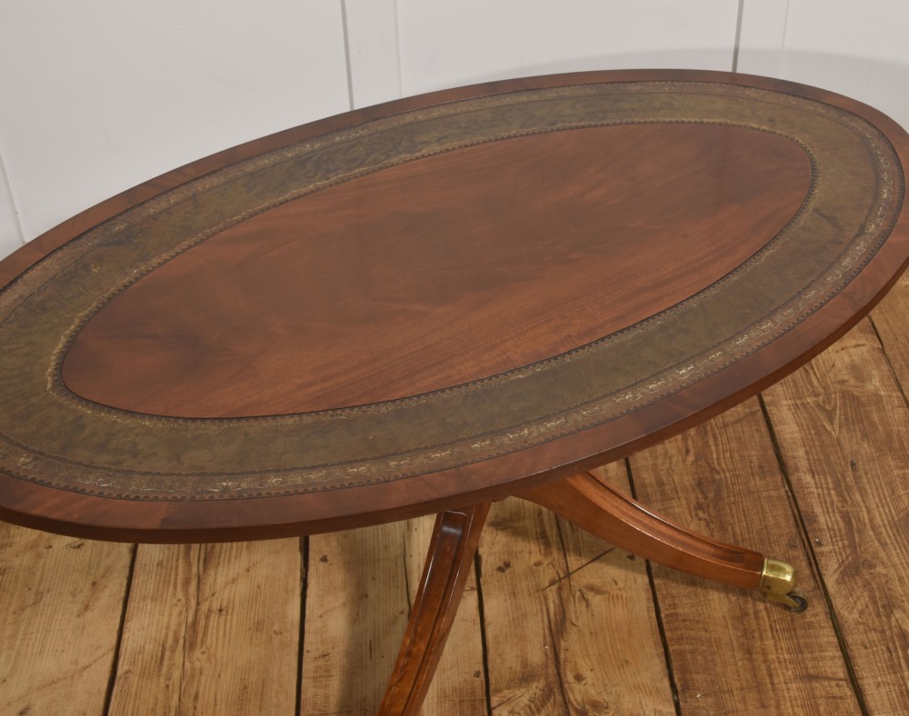 An oval mahogany coffee table featuring an inset green tooled leather border, a central turned pedestal stem, four swept legs, and brass castors. View VI
