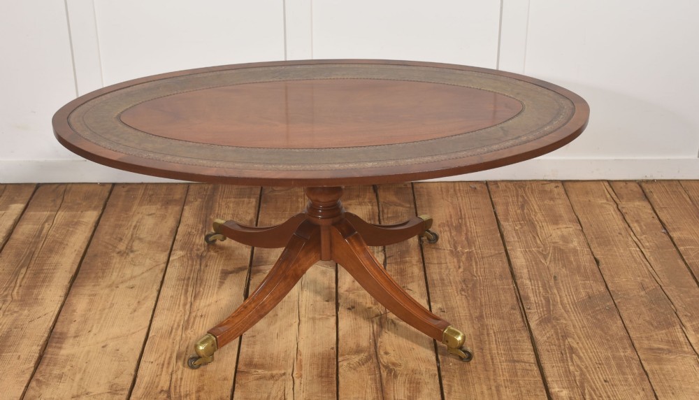An oval mahogany coffee table featuring an inset green tooled leather border, a central turned pedestal stem, four swept legs, and brass castors. View VII