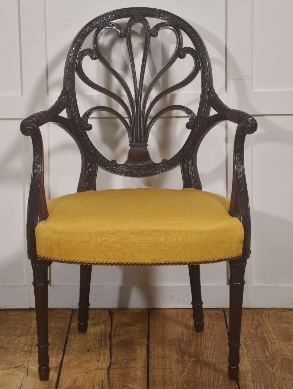 A carved mahogany open armchair featuring an oval wheat-sheaf backrest, swept arms, and a yellow upholstered seat secured with brass studs.