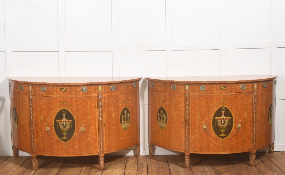 A pair of golden satinwood demi-lune cabinets featuring central hand-painted oval urn motifs and tapered legs.