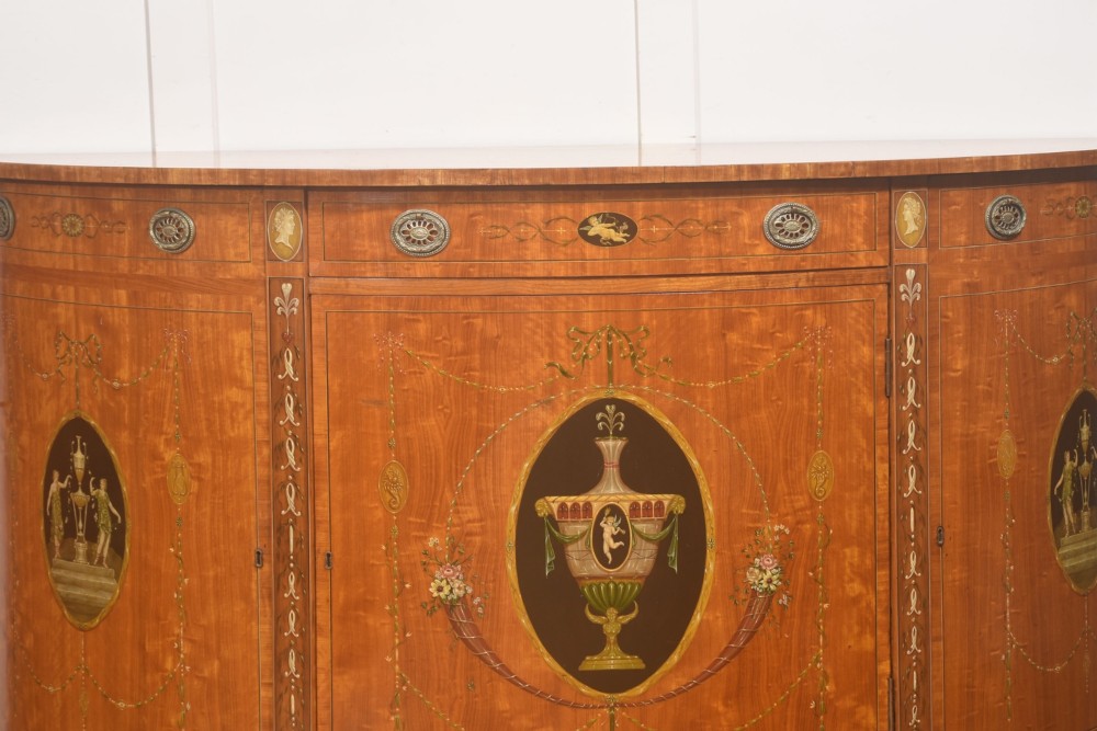 A pair of golden satinwood demi-lune cabinets featuring central hand-painted oval urn motifs and tapered legs. View VII