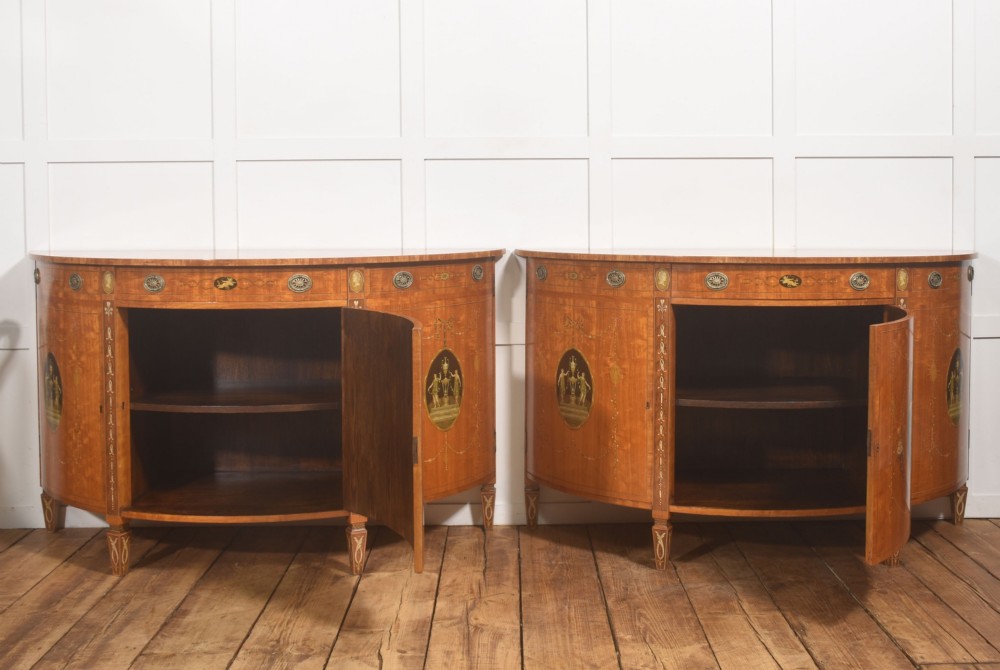 A pair of golden satinwood demi-lune cabinets featuring central hand-painted oval urn motifs and tapered legs. View X