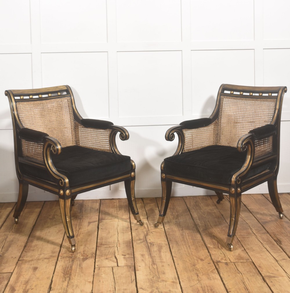 A pair of ebonised and parcel-gilt armchairs featuring woven cane backs, scroll arms, black upholstered cushions, and brass castors.