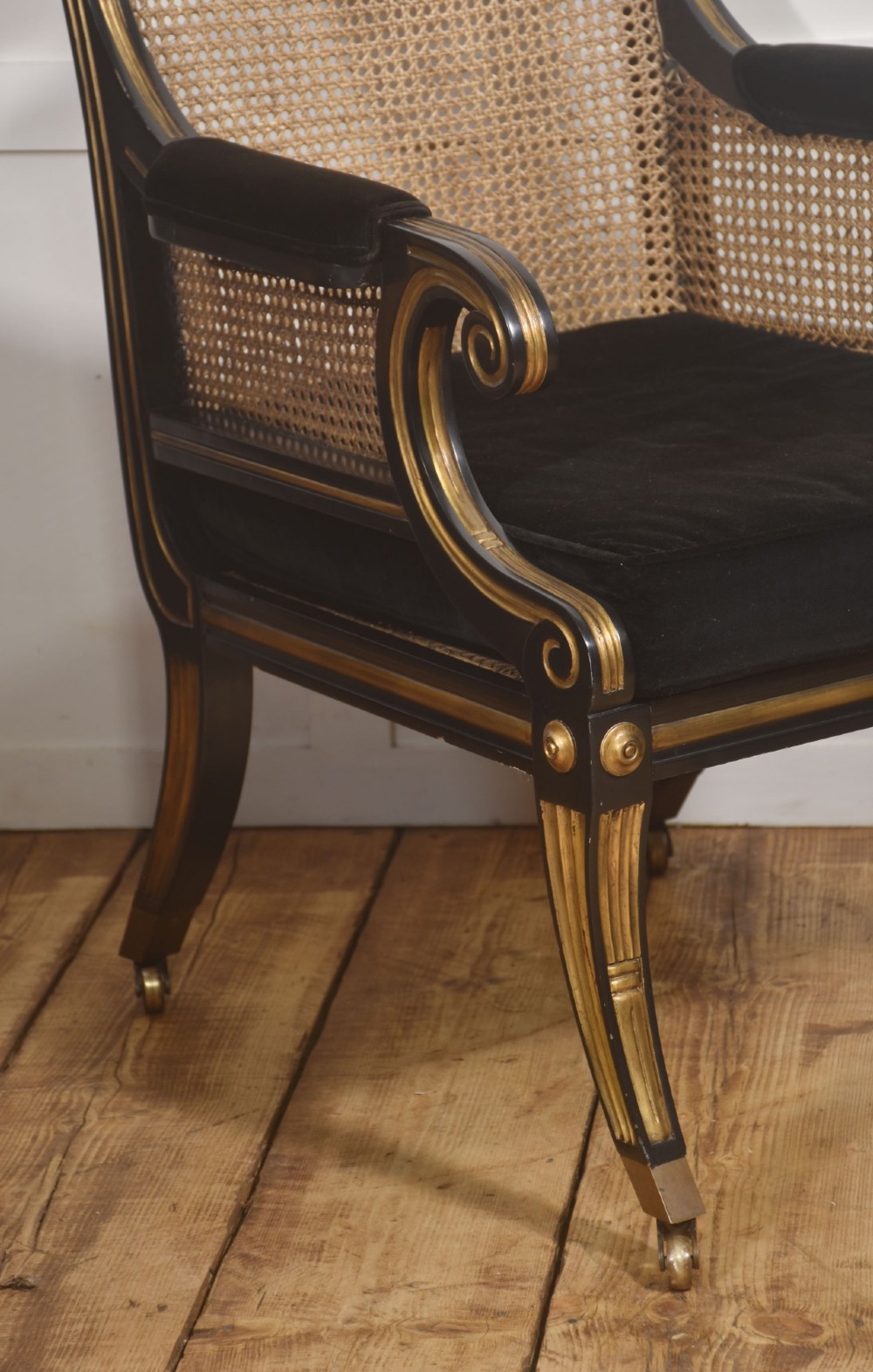 A pair of ebonised and parcel-gilt armchairs featuring woven cane backs, scroll arms, black upholstered cushions, and brass castors. View IV