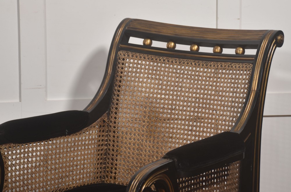 A pair of ebonised and parcel-gilt armchairs featuring woven cane backs, scroll arms, black upholstered cushions, and brass castors. View V