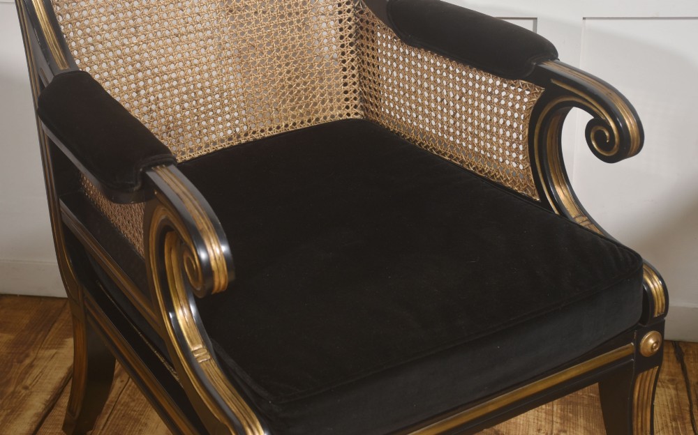 A pair of ebonised and parcel-gilt armchairs featuring woven cane backs, scroll arms, black upholstered cushions, and brass castors. View VI