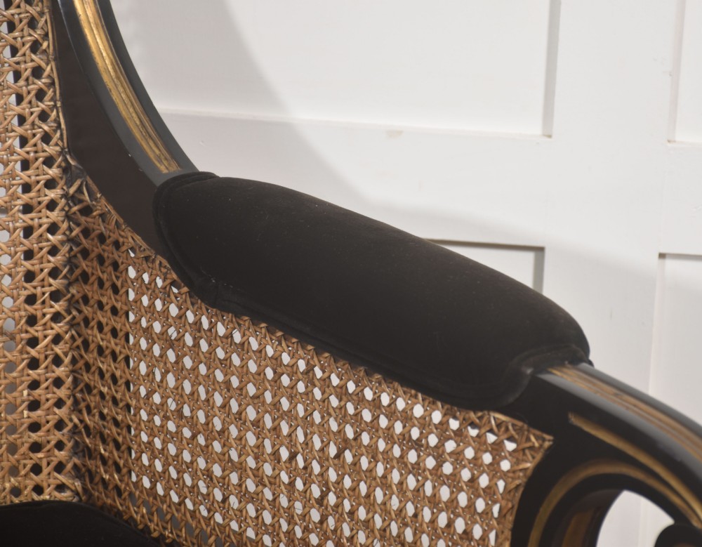 A pair of ebonised and parcel-gilt armchairs featuring woven cane backs, scroll arms, black upholstered cushions, and brass castors. View VII