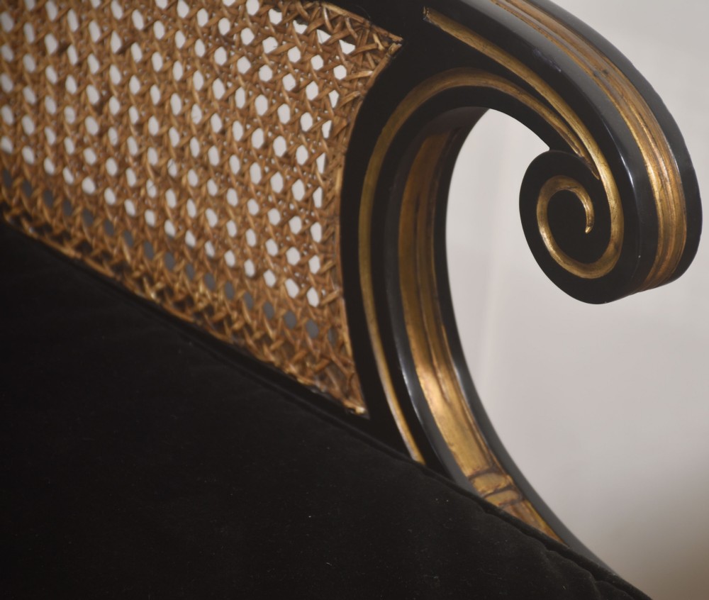 A pair of ebonised and parcel-gilt armchairs featuring woven cane backs, scroll arms, black upholstered cushions, and brass castors. View VIII