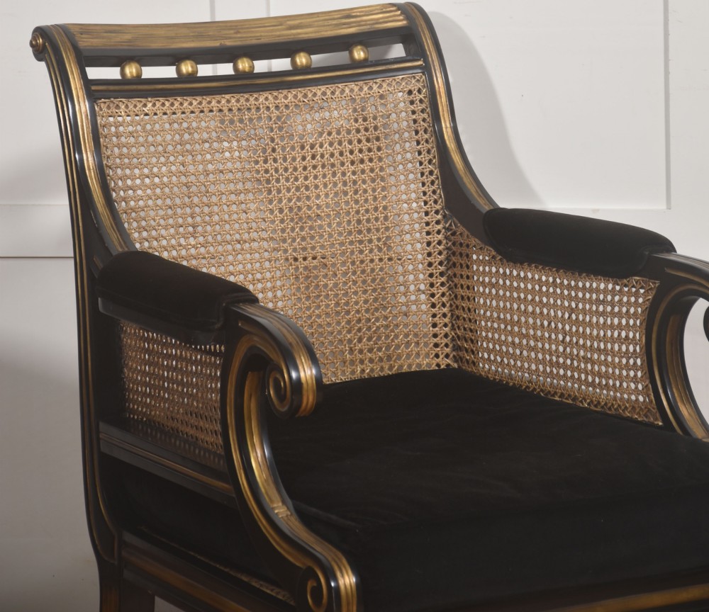 A pair of ebonised and parcel-gilt armchairs featuring woven cane backs, scroll arms, black upholstered cushions, and brass castors. View X