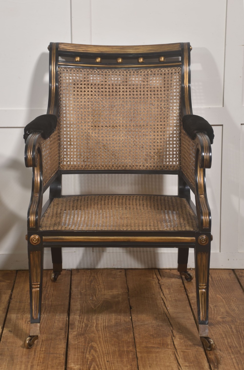 A pair of ebonised and parcel-gilt armchairs featuring woven cane backs, scroll arms, black upholstered cushions, and brass castors. View XIII