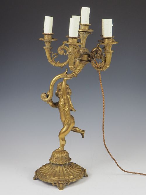 fig 1. A cast gilt brass table lamp featuring a central sculptural cherub figure supporting a six-light candelabra branch structure on an ornate base.