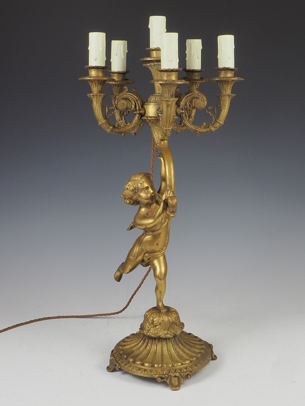 A cast gilt brass table lamp featuring a central sculptural cherub figure supporting a six-light candelabra branch structure on an ornate base.