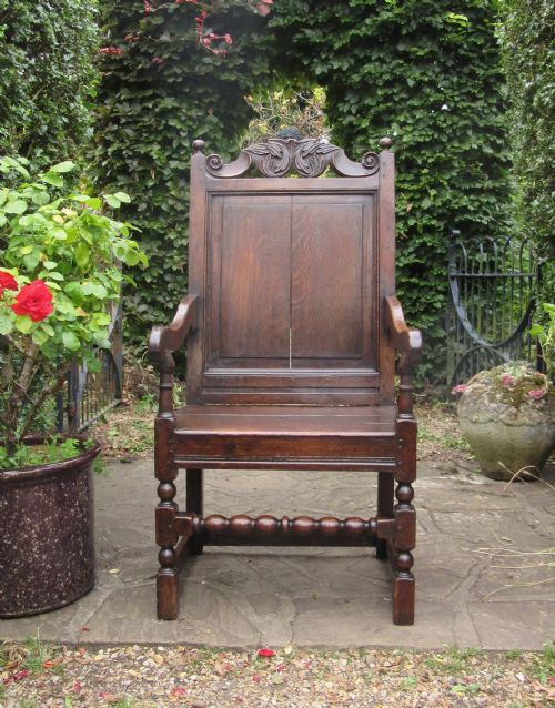 fig 1. A seventeenth-century dark oak wainscot armchair featuring a carved cresting rail, panelled back, shaped arms, and a bobbin-turned front stretcher.