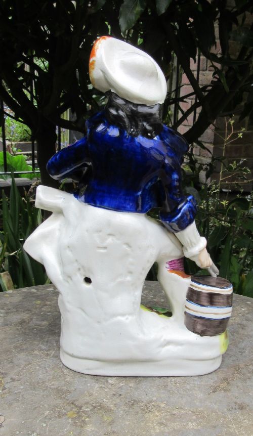 fig 2. An antique Victorian Staffordshire earthenware figurine depicting the smuggler character Will Watch seated on a stump with a barrel, wearing a cobalt blue jacket and white hat.