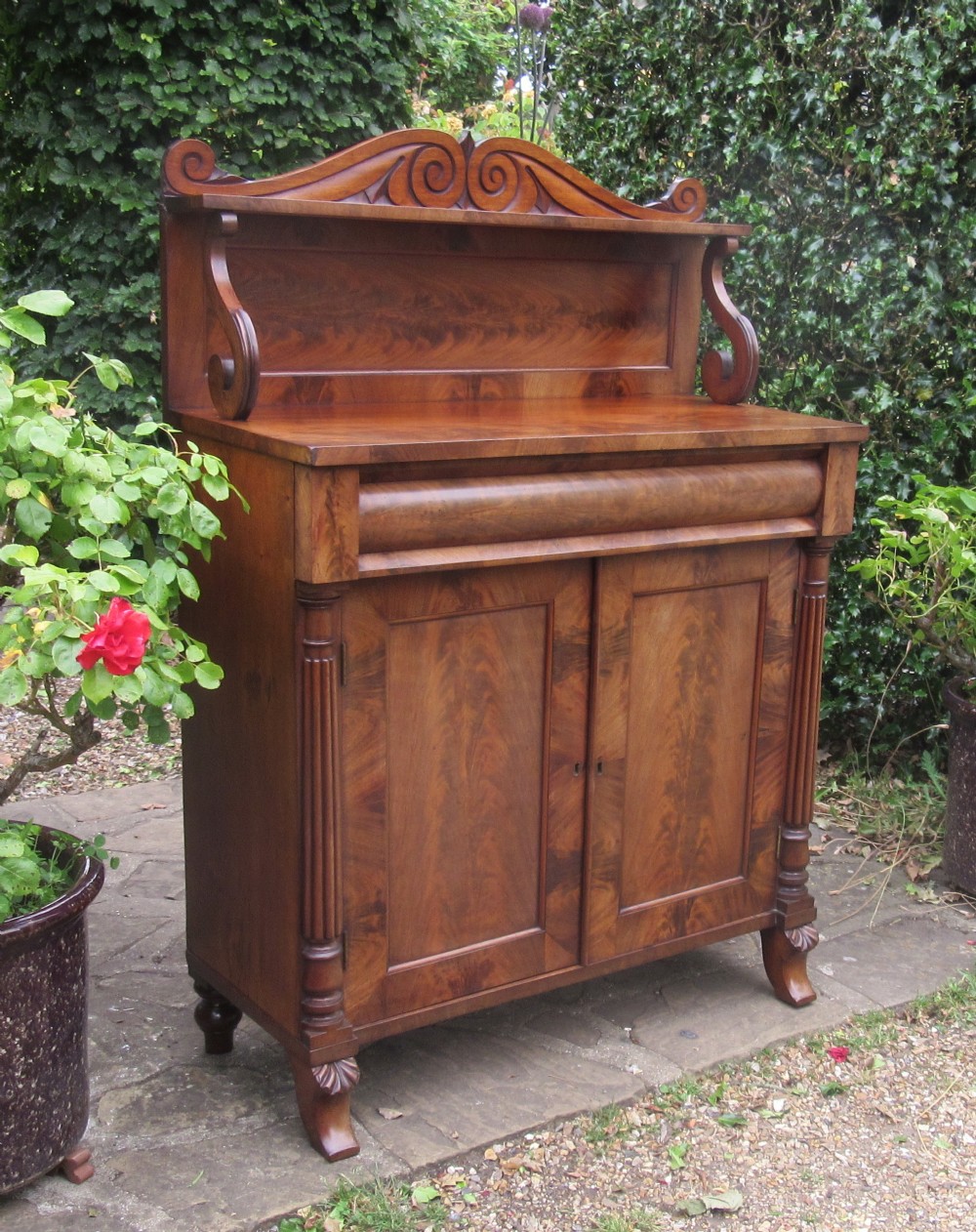 A Regency flame mahogany chiffonier featuring a carved scroll upper shelf, cushion drawer, and two cupboard doors flanked by reeded columns.