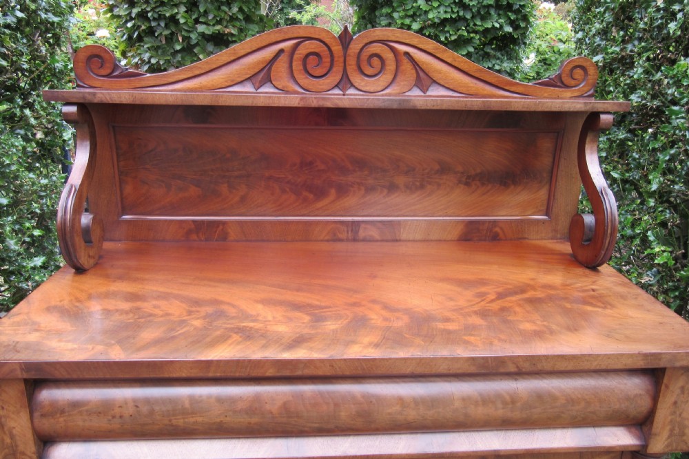 A Regency flame mahogany chiffonier featuring a carved scroll upper shelf, cushion drawer, and two cupboard doors flanked by reeded columns. View VII