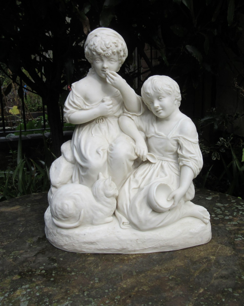 A white unglazed Parian porcelain sculpture depicting two young children sitting beside a sleeping cat on a naturalistic rock base.